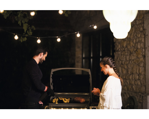 Un bărbat și o femeie fac grătar cu carne și cartofi pe un grătar exterior, luminat cu un șir de lumini.