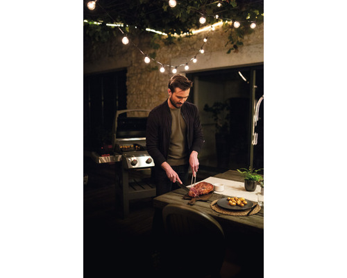 Bărbat tăind carne pe o terasă cu grătar și ghirlandă luminoasă