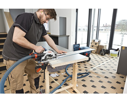 Un meșter taie o piesă de prelucrat cu un circular manual pe un banc de lucru. Un furtun de aspirare este atașat la ferăstrău.