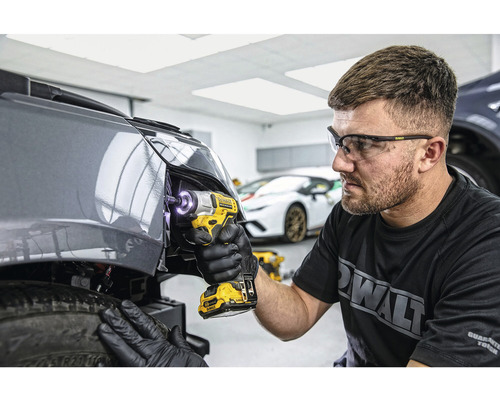 Un bărbat fixează o parte a mașinii cu o cheie de impact cu acumulator. El poartă ochelari de protecție și mănuși. În fundal se văd alte scule și mașini.