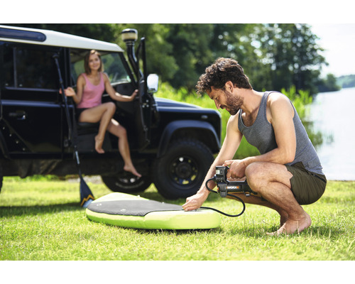 Bărbat umflă un paddleboard cu o pompă electrică, cu o femeie și un vehicul de teren în fundal.