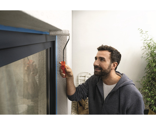 Un bărbat vopsește un cadru de fereastră cu o rolă de vopsea, care este acoperit cu bandă adezivă.