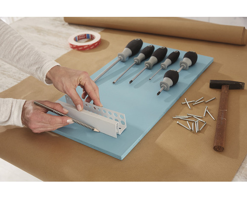 Scenă de meșteșug cu șurubelnițe, ciocan, cuie, bandă adezivă și o persoană care marchează un profil cu un creion, pe un blat de lucru.