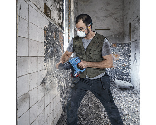 Un meseriaș în haine de lucru, cu ochelari de protecție și mască de praf, folosește un ciocan rotopercutor pe un perete.