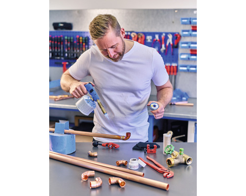 Un bărbat lipsește țevi de cupru cu un arzător cu gaz și cositor pe un banc de lucru cu unelte și fitinguri.