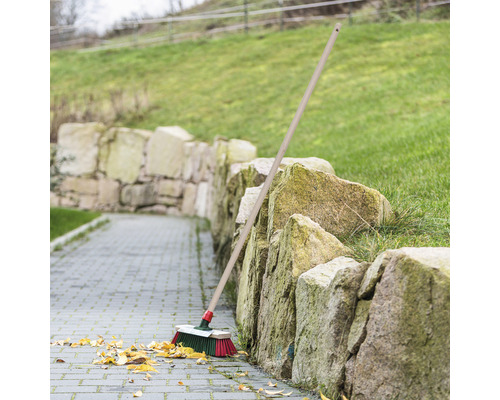 Mătură cu coadă de lemn sprijinită de un zid de piatră pe o potecă pavată cu frunze