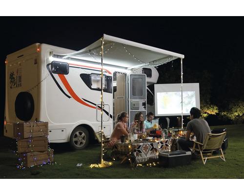 Scenă în aer liber cu rulotă, copertină, ghirlande luminoase și persoane care se uită la un ecran