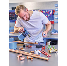 Un bărbat sudează țevi de cupru cu un arzător într-un atelier. Pe masă sunt țevi de cupru, fitinguri și unelte.