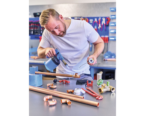 Un bărbat sudează țevi de cupru cu un arzător într-un atelier. Pe masă sunt țevi de cupru, fitinguri și unelte.