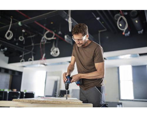 Un bărbat găurește o scândură de lemn cu o mașină de găurit. El poartă ochelari de protecție.