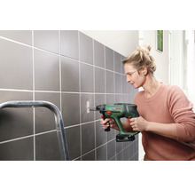 O femeie găurește un perete cu gresie cu o mașină de găurit. Poartă ochelari de protecție și dopuri pentru urechi.