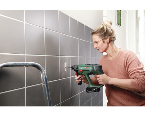 O femeie găurește un perete cu gresie cu o mașină de găurit. Poartă ochelari de protecție și dopuri pentru urechi.