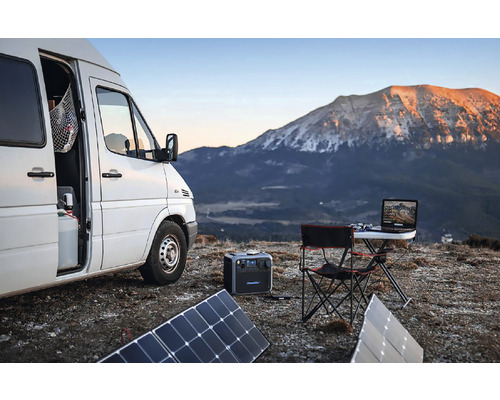 Scenă cu o furgonetă, panouri solare, o centrală electrică portabilă, o masă pliabilă și un laptop pe fundalul unui munte
