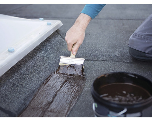 Un meșter aplică cu o pensulă un strat de acoperire pe un acoperiș plat. În prim-plan se vede o găleată cu stratul de acoperire.