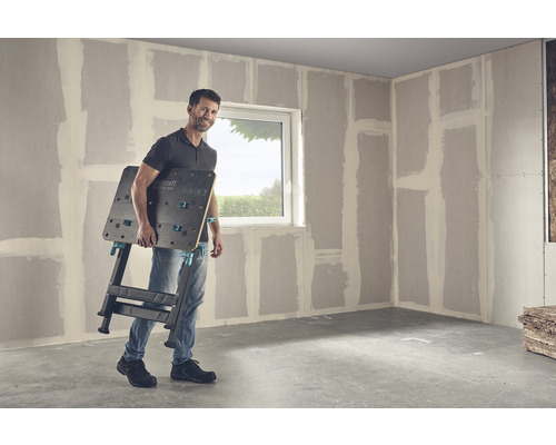 Un bărbat poartă un banc de lucru Wolfcraft Master 650 într-o cameră cu pereți din gips-carton