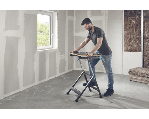 Bărbat lucrând cu un banc de lucru într-o cameră cu pereți din gips-carton și o fereastră.