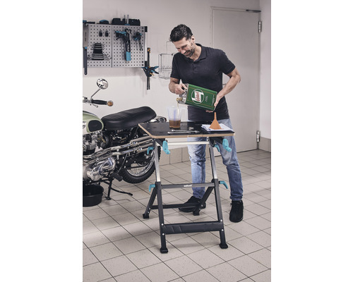 Bărbat turnând ulei pe bancul de lucru într-un garaj, cu o motocicletă și un perete cu scule în fundal.