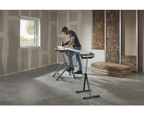 Un bărbat lucrează cu un ferăstrău vertical pe o piesă de lucru pe un banc de lucru Wolfcraft într-o cameră cu plăci de gips-carton și material izolant.