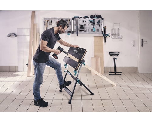 Un bărbat găurește o piesă de lucru cu o mașină de găurit cu acumulator pe un banc de lucru Wolfcraft Master 600.