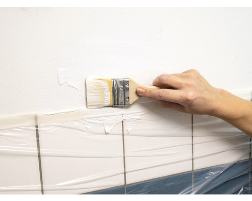 O persoană vopsește cu o pensulă un perete deasupra faianței acoperite cu folie.