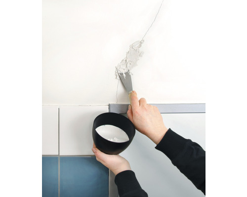 O persoană astupă o fisură în perete cu o spatulă. Se ține un vas cu material de umplere.
