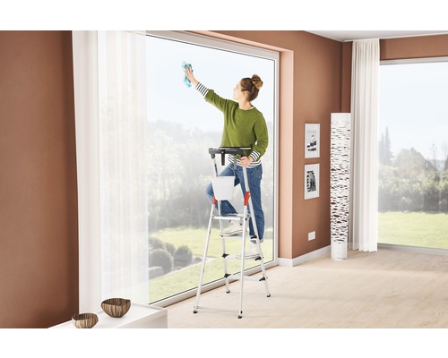Femeie curățând ferestrele stând pe o scară cu găleată și suport într-o cameră de zi.
