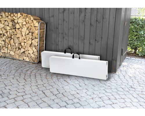 Două mese pliante albe pliate cu mânere de transport stau afară în fața unui șopron de lemn.