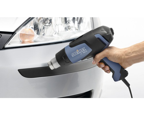 Mână ținând pistolul cu aer cald STEINEL HL 1820 S în fața unei bare de protecție argintii