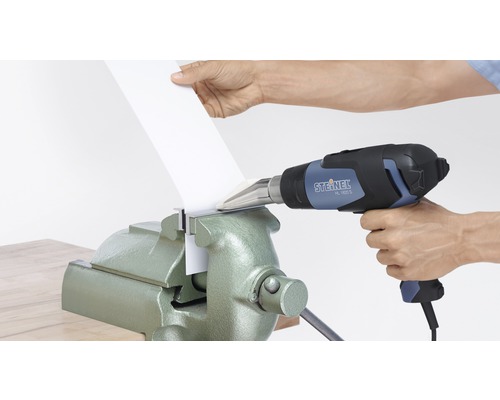 O persoană încălzește o piesă de prelucrat cu un pistol cu aer cald, care este fixată într-o menghină.