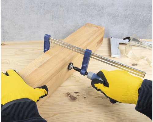 O șipcă de lemn este fixată cu o menghină cu șurub, acționată de mâini cu mănuși de lucru.