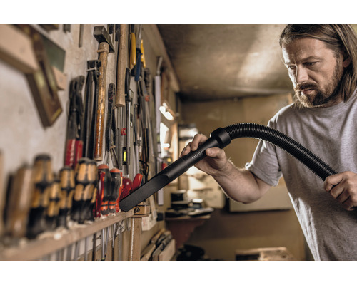 Artizan curățând bancul de lucru cu un cap de aspirator în fața unui perete cu scule