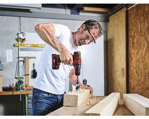 Un bărbat găurește o scândură de lemn cu o bormașină cu acumulator. Poartă ochelari de protecție.