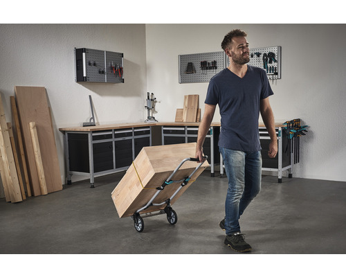 Un bărbat transportă lemn cu un cărucior de transport într-un atelier cu banc de lucru și perete pentru scule