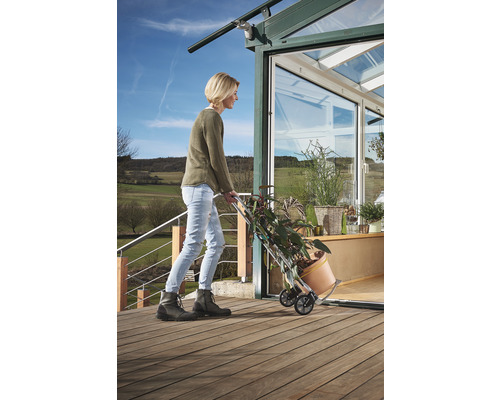 Femeie transportând plante cu un cărucior în fața unei sere.