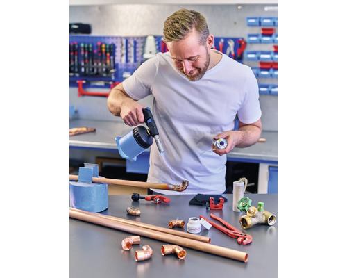 Bărbat care sudează țevi de cupru cu un arzător cu gaz și cositor pe un banc de lucru.