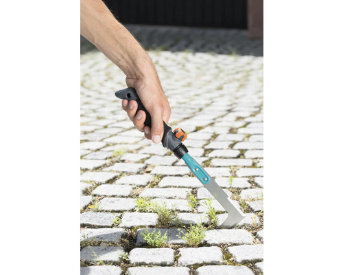 O mână ține o racletă pentru rosturi pentru a îndepărta buruienile dintre pietrele de pavaj.