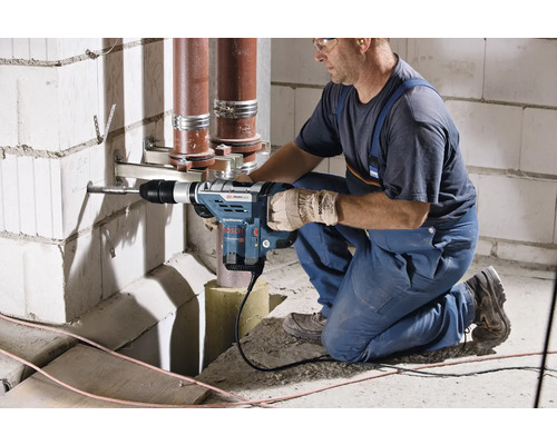 Un meșter găurește un perete cu un ciocan rotopercutor, conducte care trec în fundal.