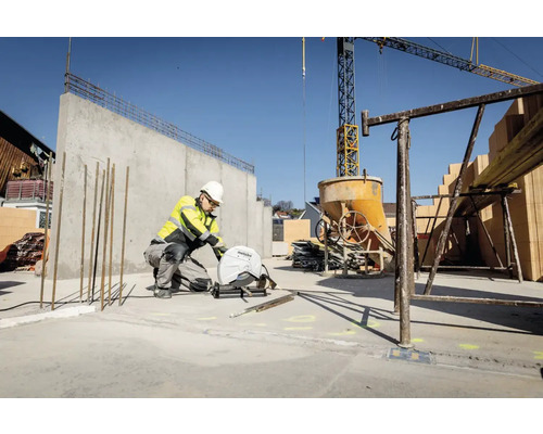 Un muncitor în construcții, în echipament de protecție, taie oțel cu un ferăstrău Metabo pe un șantier, cu pereți de beton și o macara în fundal.