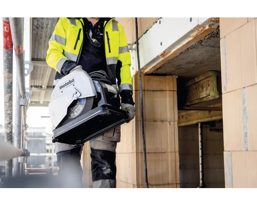 Muncitor în construcții care transportă un ferăstrău metalic Metabo pe un șantier.