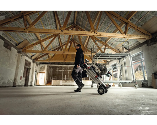 Un bărbat transportă o schelă mobilă pliată într-o încăpere cu tavan cu grinzi de lemn.