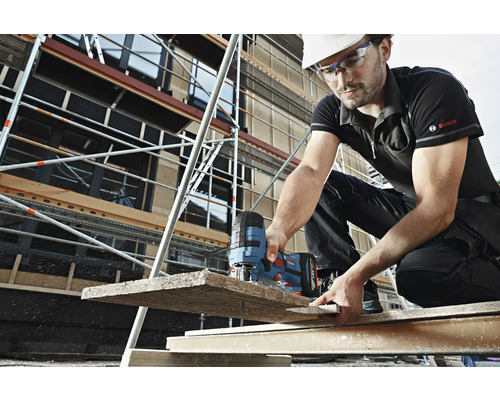 Un muncitor în construcții cu cască și ochelari de protecție taie o scândură de lemn cu un ferăstrău vertical pe un șantier. În fundal se vede o schelă. Logo Bosch.