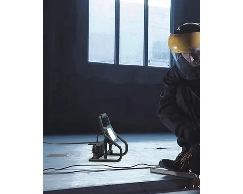 Muncitor care prelucrează metal folosind un reflector de șantier pentru a ilumina zona de lucru.