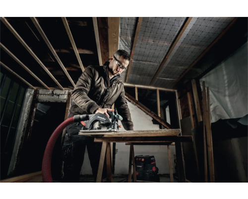 Artizan cu ochelari de protecție tăind lemn cu un ferăstrău circular portabil Metabo într-o clădire în construcție.