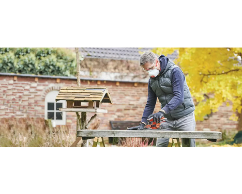 Un bărbat șlefuiește o scândură de lemn în aer liber cu o șlefuitoare electrică, purtând o mască de protecție și ochelari de protecție.