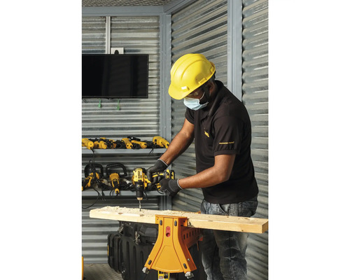 Un bărbat găurește o scândură de lemn așezată pe un banc de lucru cu o bormașină. În fundal se văd alte bormașini și unelte.