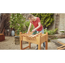 Femeie șlefuind lemn pe un strat de grădină înălțat cu o șlefuitoare în grădină.