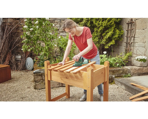 Femeie șlefuind lemn pe un strat de grădină înălțat cu o șlefuitoare în grădină.