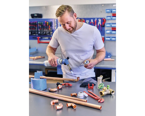 Bărbat care sudează țevi de cupru cu un arzător cu gaz, înconjurat de fitinguri de țevi de cupru și scule