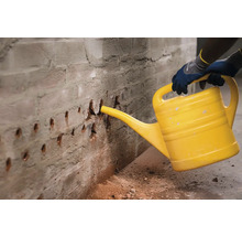 O persoană udă un perete de cărămidă cu o stropitoare galbenă pentru a lega praful.