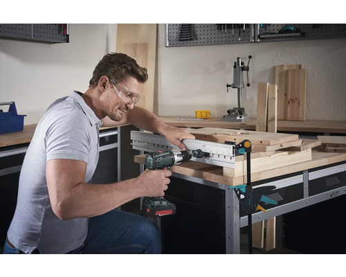 Un bărbat găurește cu o mașină de găurit o placă de lemn cu șablon de găurire pe un banc de lucru.